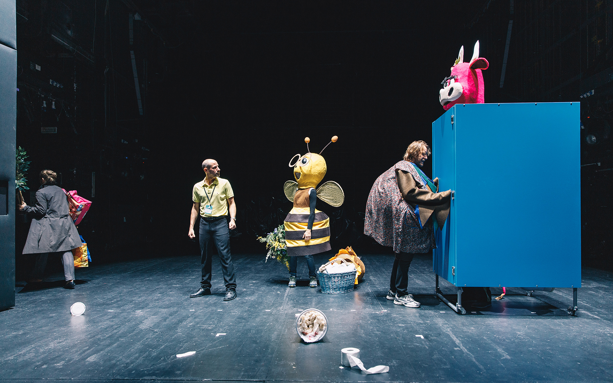 Igralec, kostumiran v velikega ptiča (Matej Puc), je snel zgornji del kostuma in se pogovarja z igralcem, kostumiranim v veliko roza kravo (Lotos Vincenc Šparovec), ki sedi na stranišču, v ozadju se igralka, kostumirana v veliko čebelo (Mojca Funkl), pogo