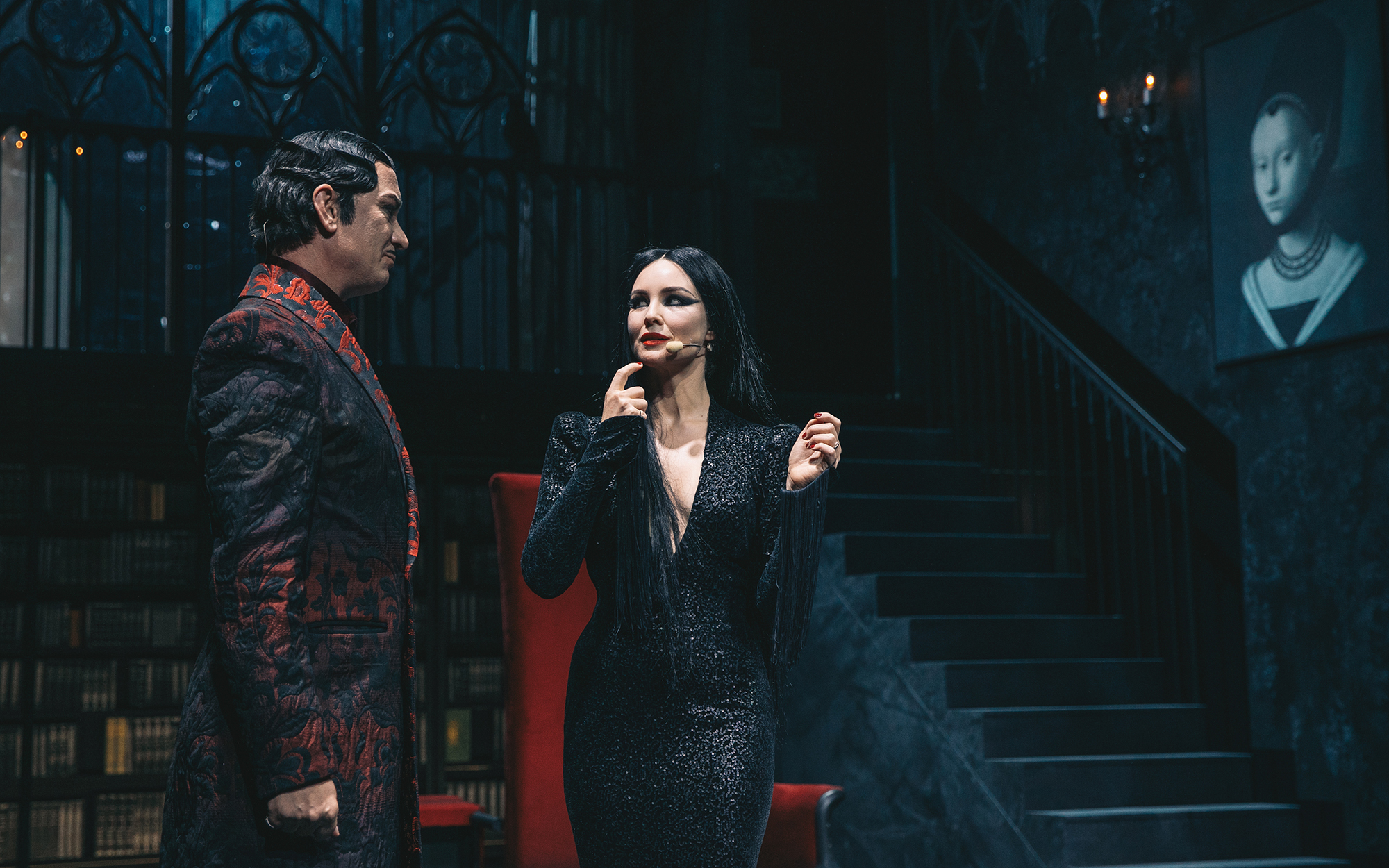 Morticija Addams (Iva Krajnc Bagola) in Gomez Addams (Uroš Smolej) stojita v dnevnem prostoru in se pogovarjata, v ozadju so stopnišče in gotska okna graščine. Avtor fotografije: Peter Giodani.