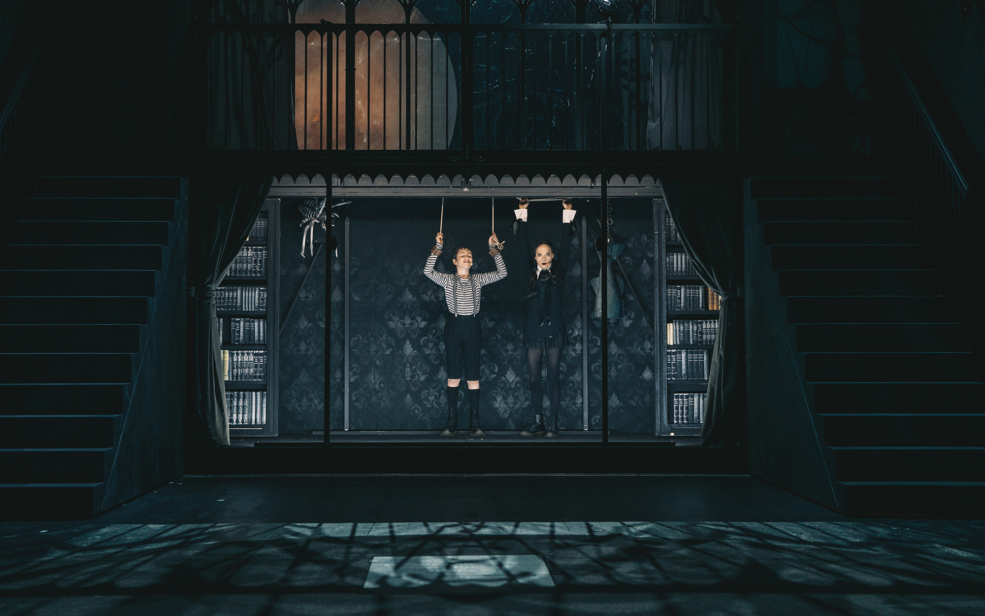 Bucko Addams (Jurij Weiss) in Sreda Addams (Viktorija Bencik Emeršič) stojita z dvignjenimi rokami v niši, ki je ravno dovolj velika zanju. Bucko Addams (Jurij Weiss) ima zavezana zapestja na strop. Avtor fotografije: Peter Giodani.