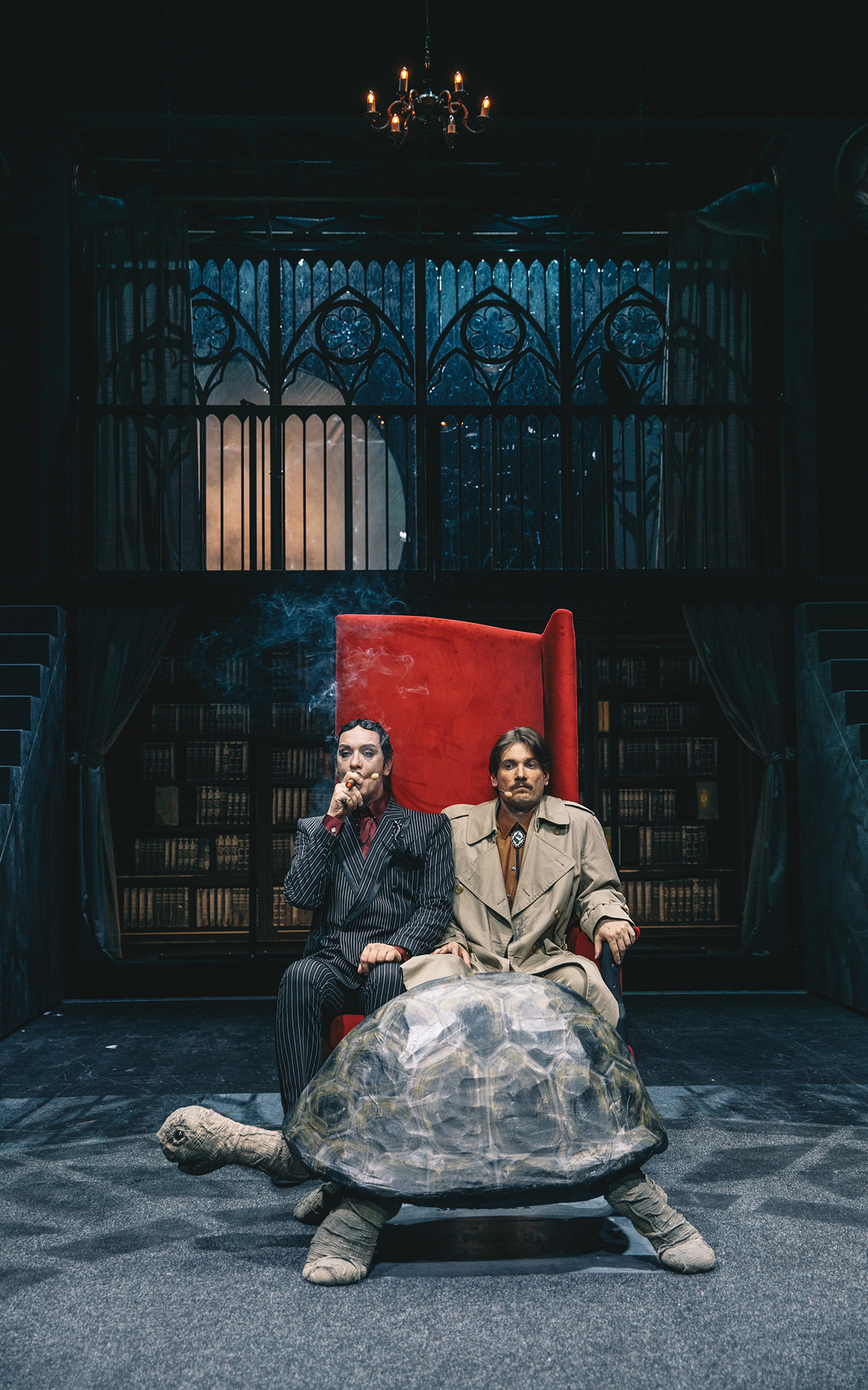 Gomez Addams (Uroš Smolej) in Jan Krneki (Robert Korošec) sedita stlačena v rdečem naslanjaču, Gomez Addams kadi cigaro. Sta na sredini dnevnega prostora, v ozadju ogromno stopnišče, nad njim vrsta gotskih oken. Avtor fotografije: Peter Giodani.