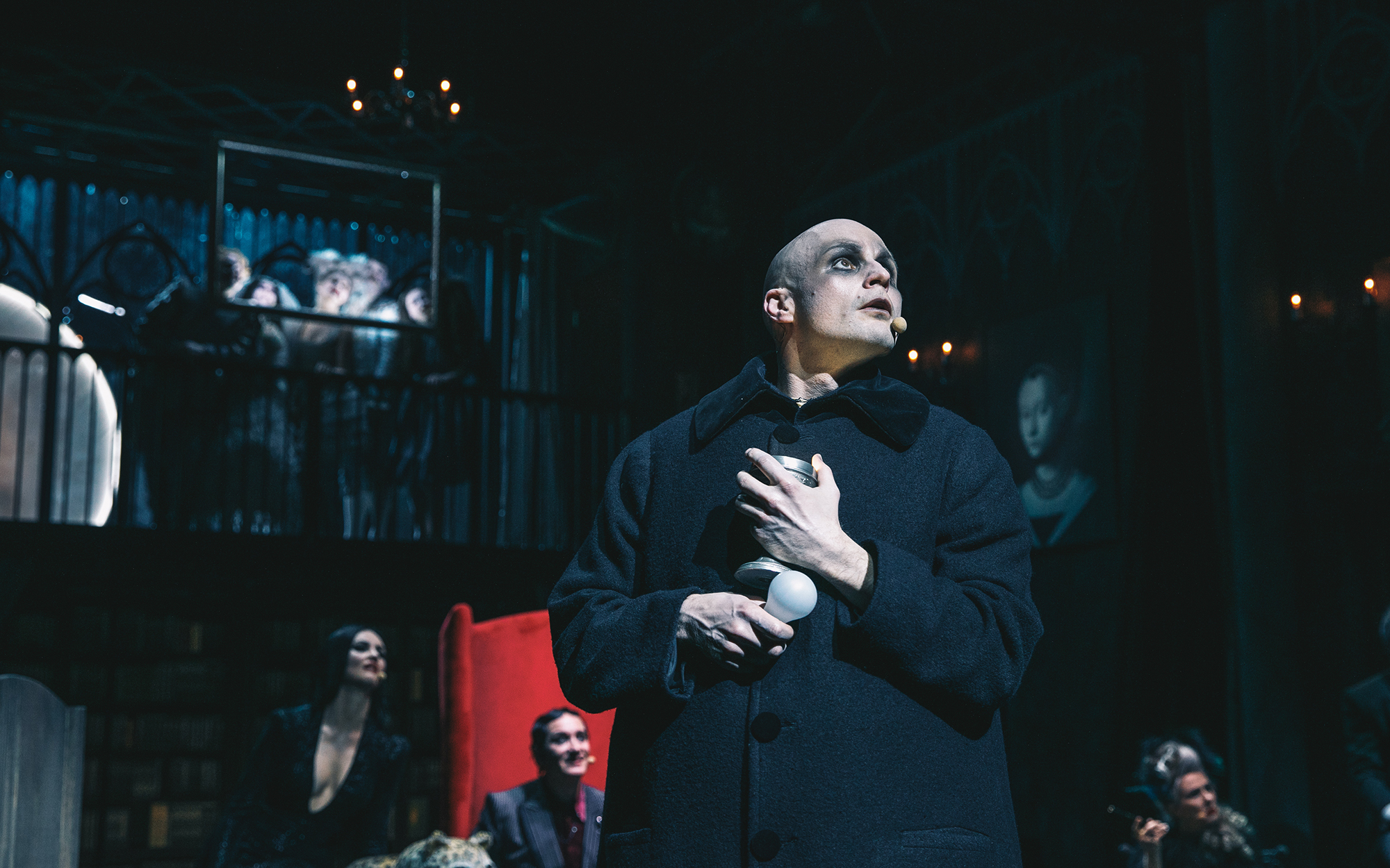 Gnoj Addams (Jaka Lah) privija k sebi kelih in žarnico ter nekaj opazuje, obrnjen je proti občinstvu. Avtor fotografije: Peter Giodani.
