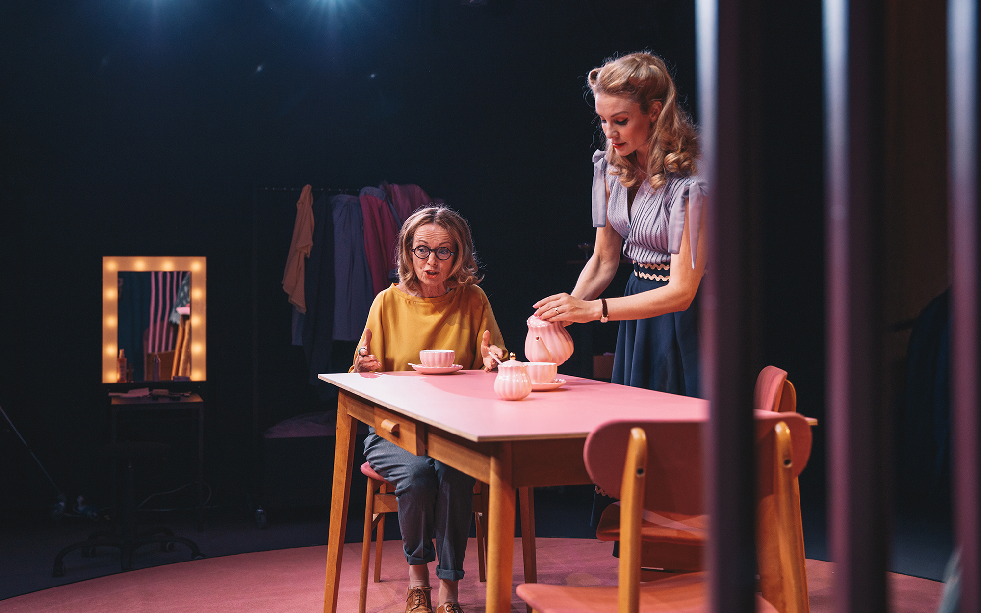 Sylvia (Bernarda Oman) nekaj vneto govori, medtem ko sedi za kuhinjsko mizo, pred njo je rožnati servis za čaj, Judy (Iva Krajnc Bagola) jo posluša in si nataka čaj. Avtor fotografije: Peter Giodani.