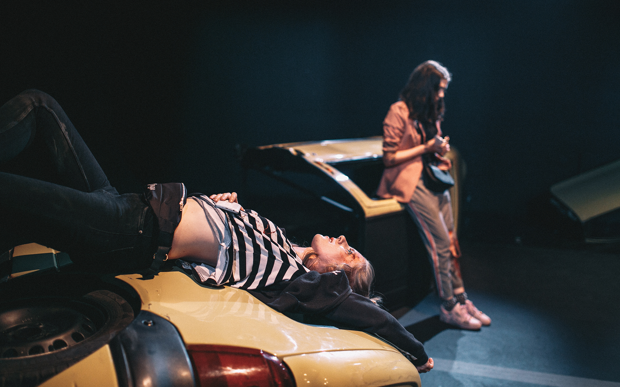 Leni (Lena Hribar Škrlec) z okrvavljenim obrazom leži na delu razbitega avtomobila in se pogovarja z Viktorijo (Anuša Kodelja), ki se naslanja na druge razbitine avtomobila. Avtor fotografije: Peter Giodani.