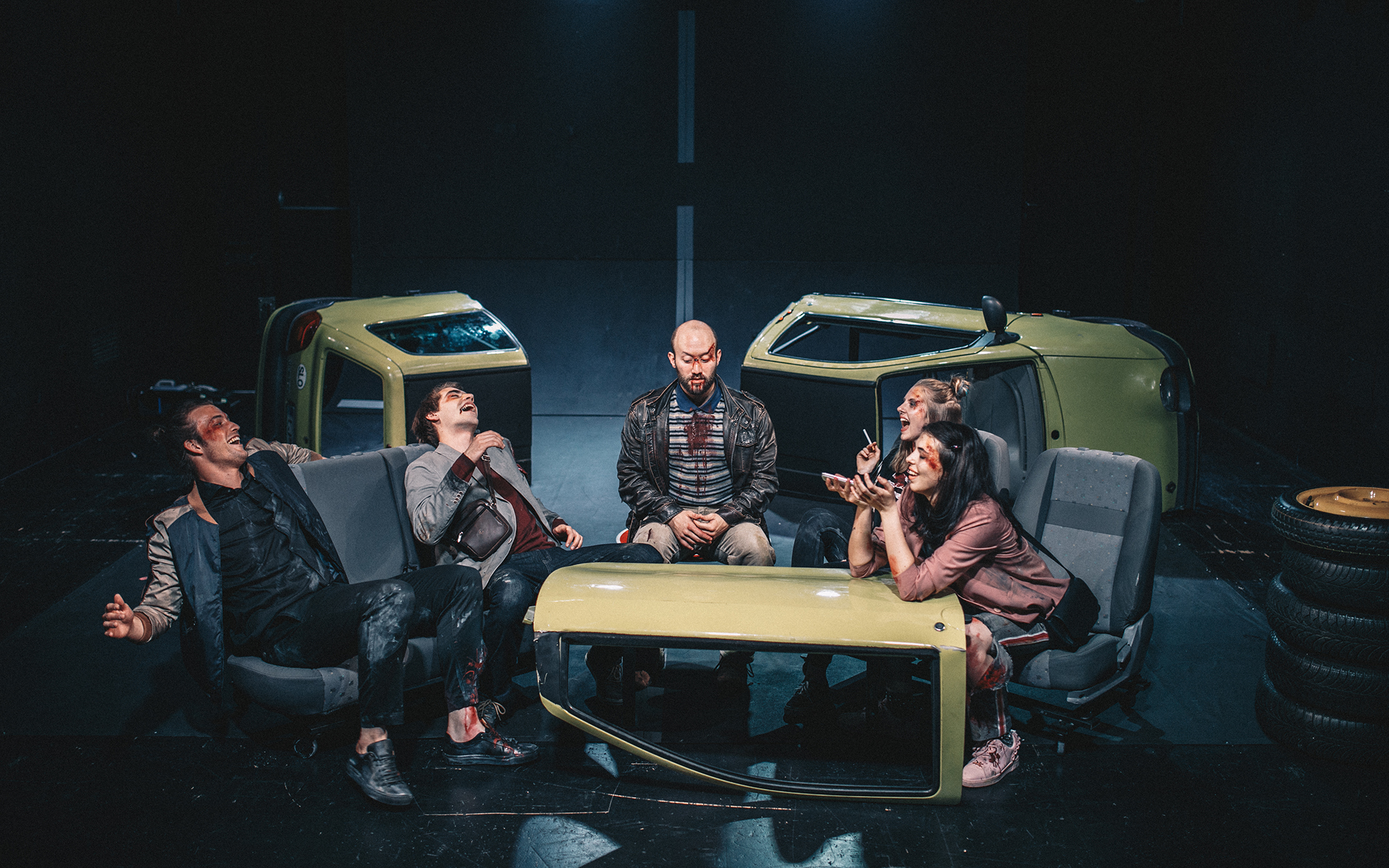 Peter (Filip Samobor), Žan (Matic Lukšič), Dejan (Tines Špik), Leni (Lena Hribar Škrlec) in Viktorija (Anuša Kodelja) nasmejani sedijo na razbitinah avtomobila; iz enega dela so si naredili provizorično mizico, Viktorija se s komolci naslanja nanjo. Avtor