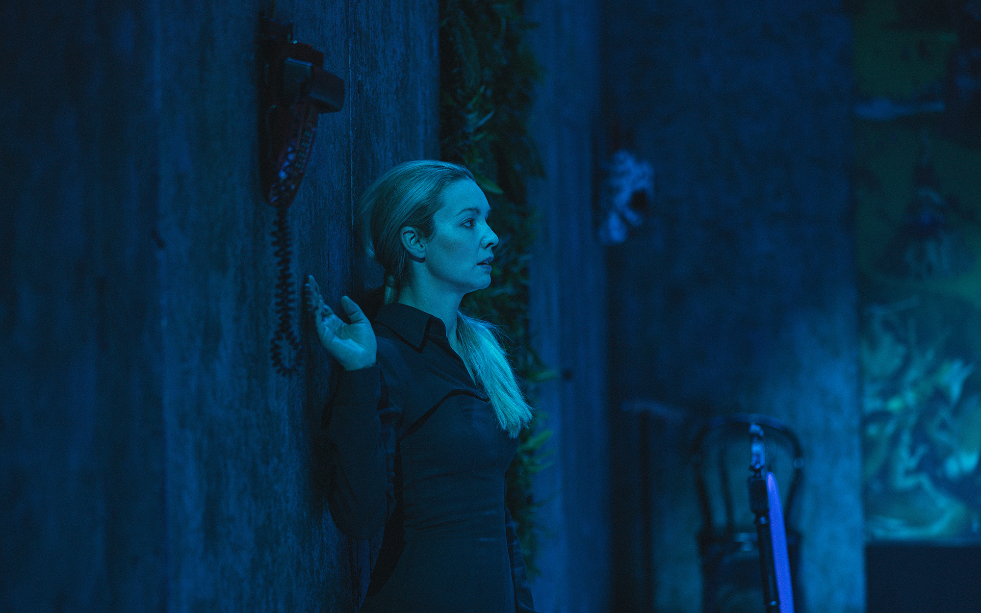 Ženska (Iva Krajnc Bagola) je s hrbtom naslonjena ob steno, z roko prestrašeno sega po stenskem telefonu. Na desni strani na steni visi ena od živalskih mask. Sobo obdaja hladna modra svetloba.