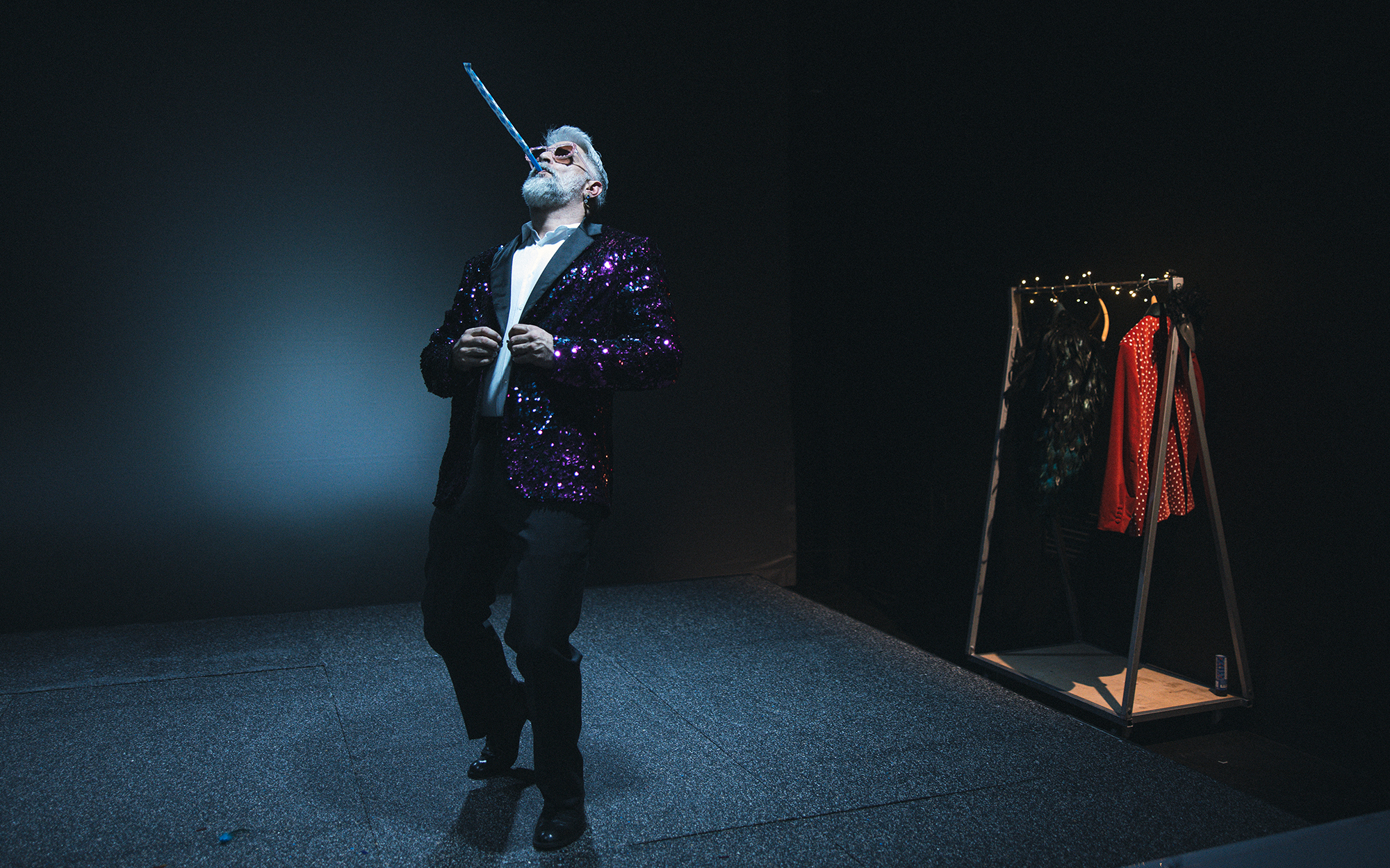 On (Sebastian Cavazza) v plesnem koraku piha v papirnato piščal, ki jo uporabljamo ob praznovanjih. Ob tem si zapenja bleščeč suknjič. Avtor fotografije: Peter Giodani.
