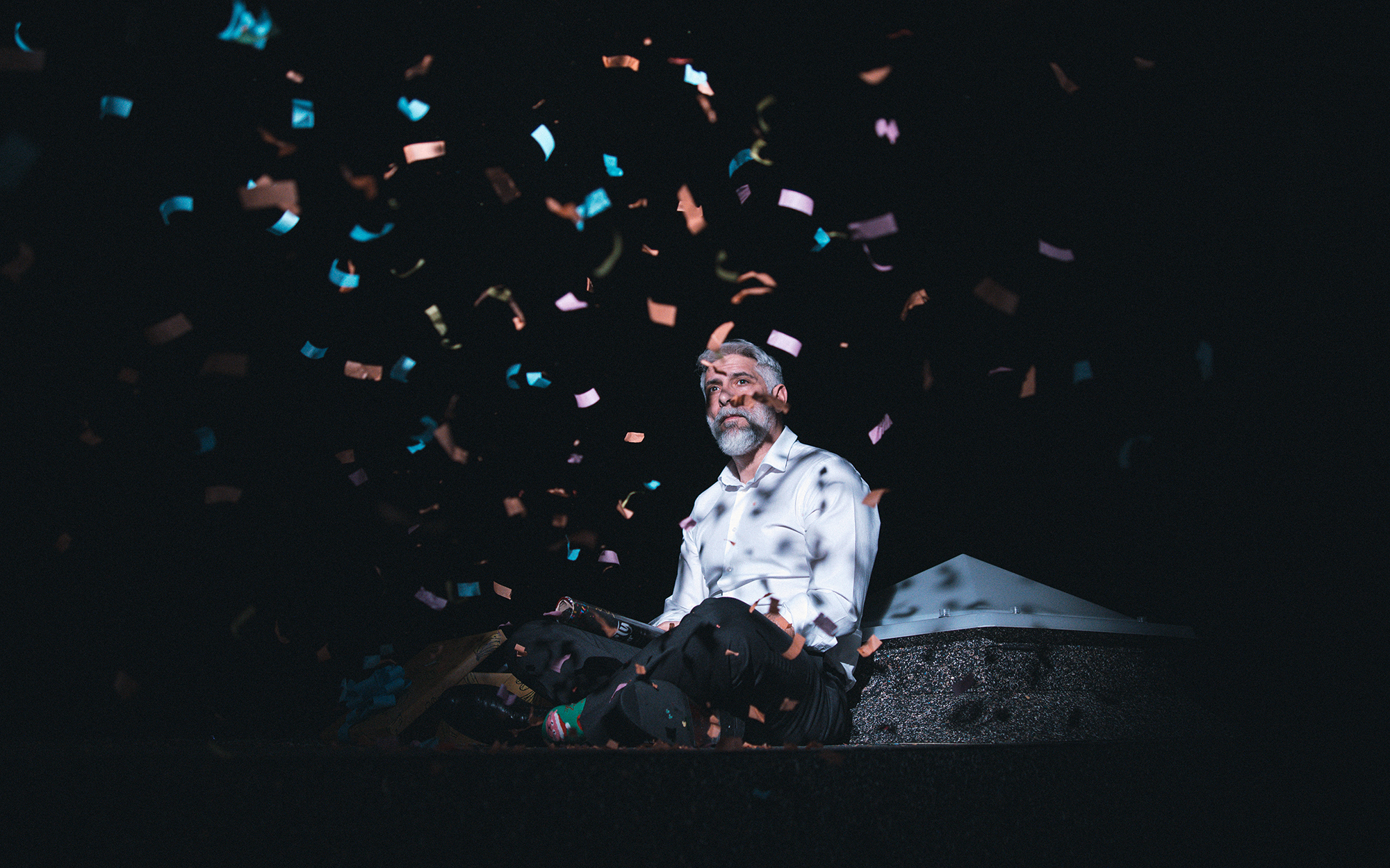 On (Sebastian Cavazza) s prekrižanimi nogami sedi naslonjen na strešno svetlobno kupolo in opazuje konfete, ki se usipajo nanj. Avtor fotografije: Peter Giodani.