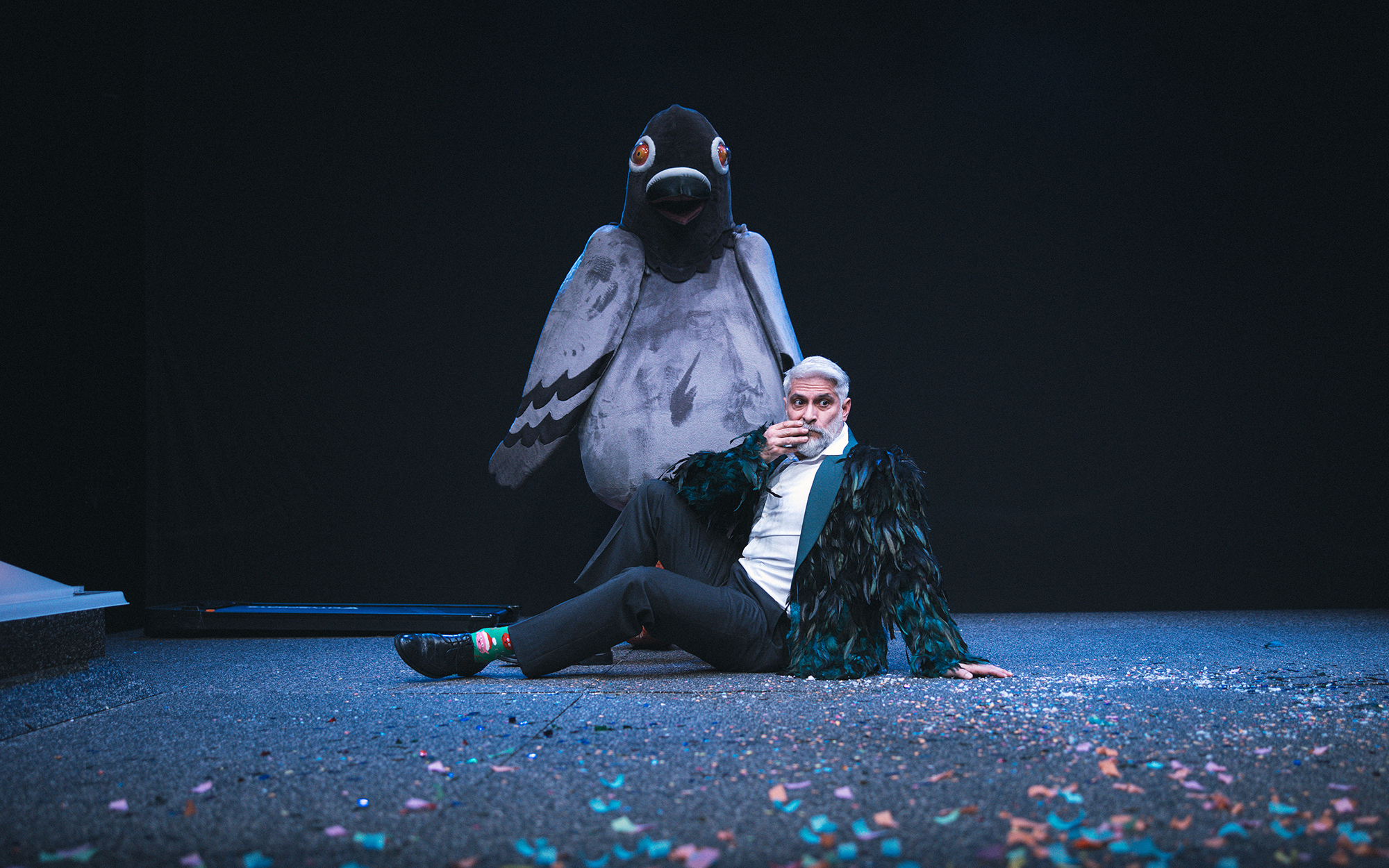 On (Sebastian Cavazza) sedi na tleh, oblečen je v sako s ptičjimi peresi. Za njim stoji Ona (Ajda Smrekar), kostumirana v velikega goloba. Avtor fotografije: Peter Giodani.