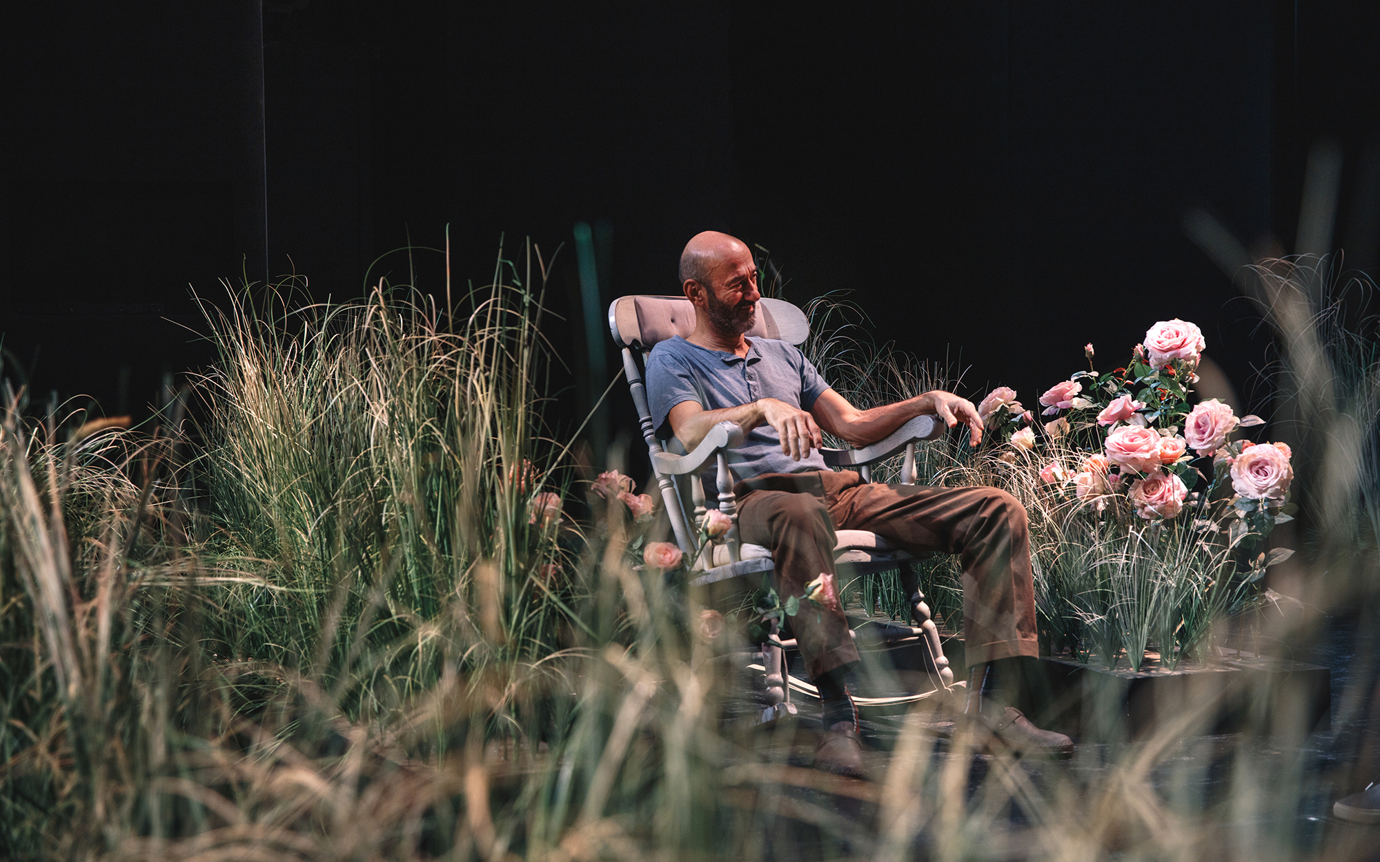 Bob (Boris Ostan) zamišljeno sedi v gugalniku na vrtu, obdajajo ga visoka trava in vrtnice. Avtor fotografije: Peter Giodani.