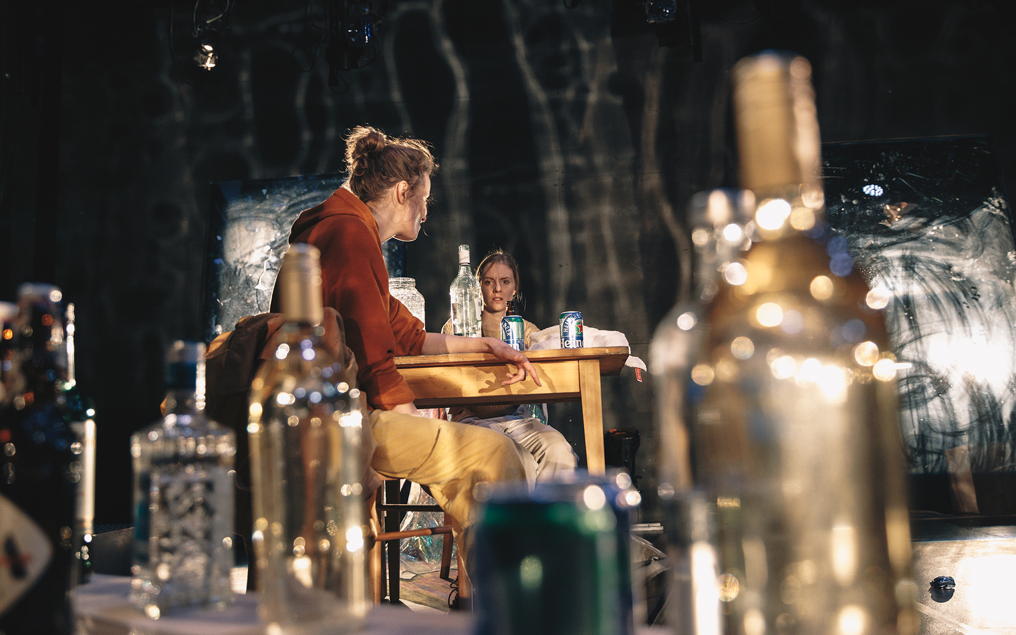 Od strani opazujemo Jess (Ajda Smrekar) in Sarah (Lena Hribar Škrlec), ki sedita za kuhinjsko mizo, pijeta in se pogovarjata. Na mizi kozarec z ribo, steklenica in dve pločevinki piva. V ospredju je na tleh več steklenic. Avtor fotografije: Peter Giodani.