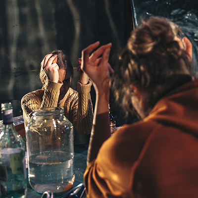 Sarah (Lena Hribar Škrlec) in Jess (Ajda Smrekar) sta s komolci naslonjeni na kuhinjsko mizo, na kateri je kozarec z ribo. Tleskata s prsti in se gledata prek mize. Okoli njiju so steklenice in pločevinke. Avtor fotografije: Peter Giodani.