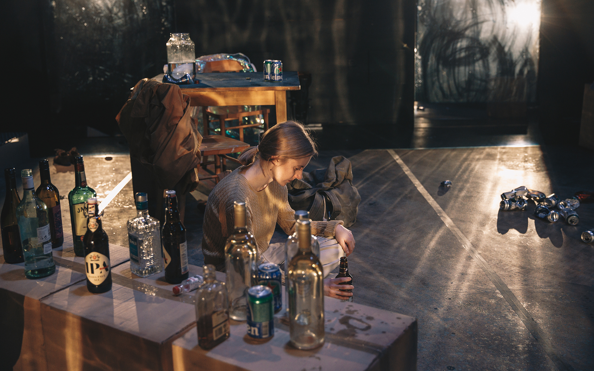 Sarah (Lena Hribar Škrlec) sama sedi ob kartonastih škatlah, na katerih so steklenice in pločevinke alkohola. V roki drži steklenico s pivom. V ozadju je kuhinjska miza, na kateri je kozarec z ribo. Avtor fotografije: Peter Giodani.