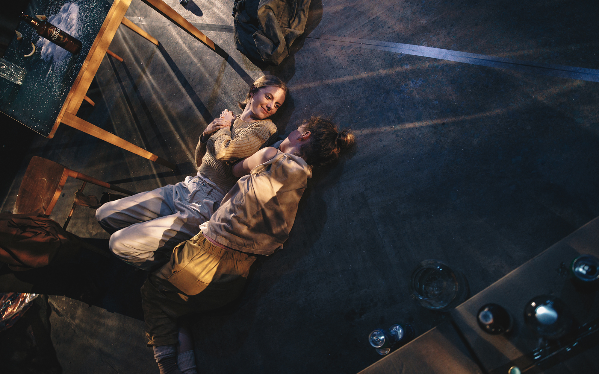 Jess (Ajda Smrekar) in Sarah (Lena Hribar Škrlec) ležita na tleh ob kuhinjski mizi ter se gledata in smehljata. Avtor fotografije: Peter Giodani.