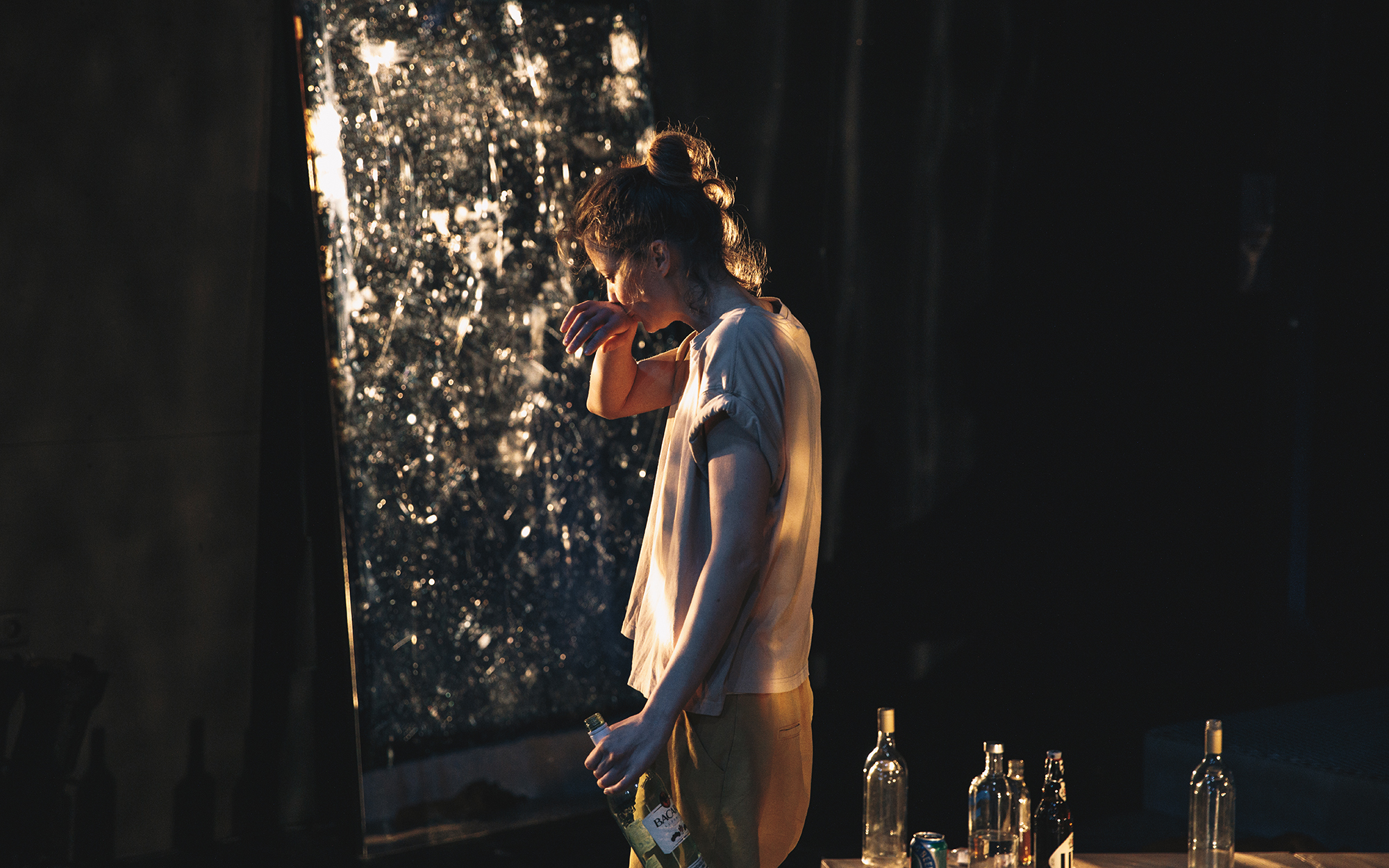 Jess (Ajda Smrekar) stoji sama s steklenico v eni roki, z drugo si briše usta. Avtor fotografije: Peter Giodani. 