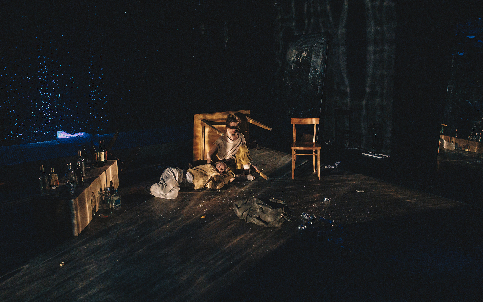 V zavetju prevrnjene mize sedi Jess (Ajda Smrekar) in boža spečo Sarah (Lena Hribar Škrlec), ki leži ob njej. Avtor fotografije: Peter Giodani.