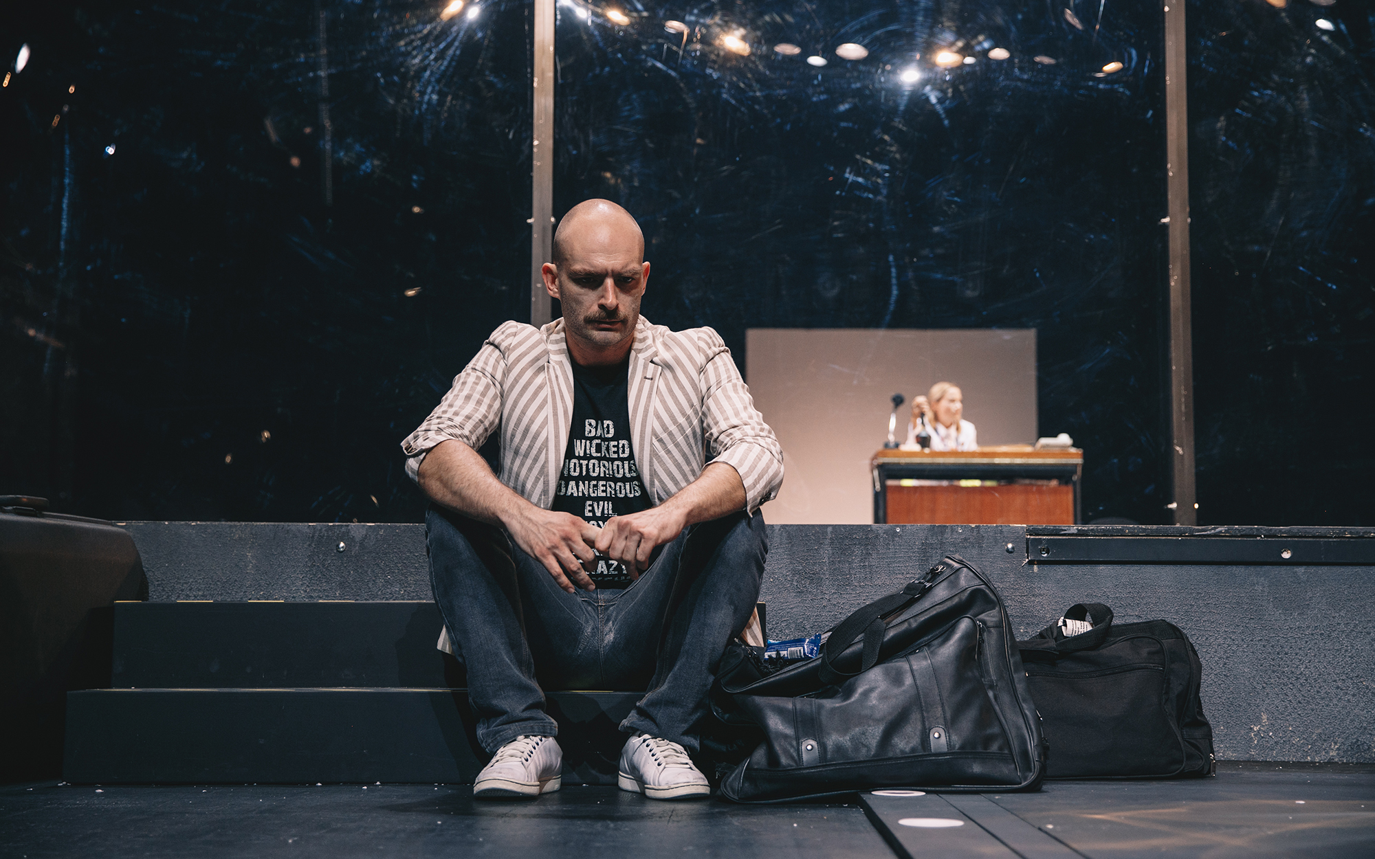 Toni (Gregor Gruden) sedi na stopnicah in zamišljeno gleda predse, ob njem sta dva kosa prtljage. V ozadju za stekleno steno je silhueta Cherie (Viktorija Bencik Emeršič), ki sedi za pisalno mizo. Avtor fotografije: Peter Giodani.