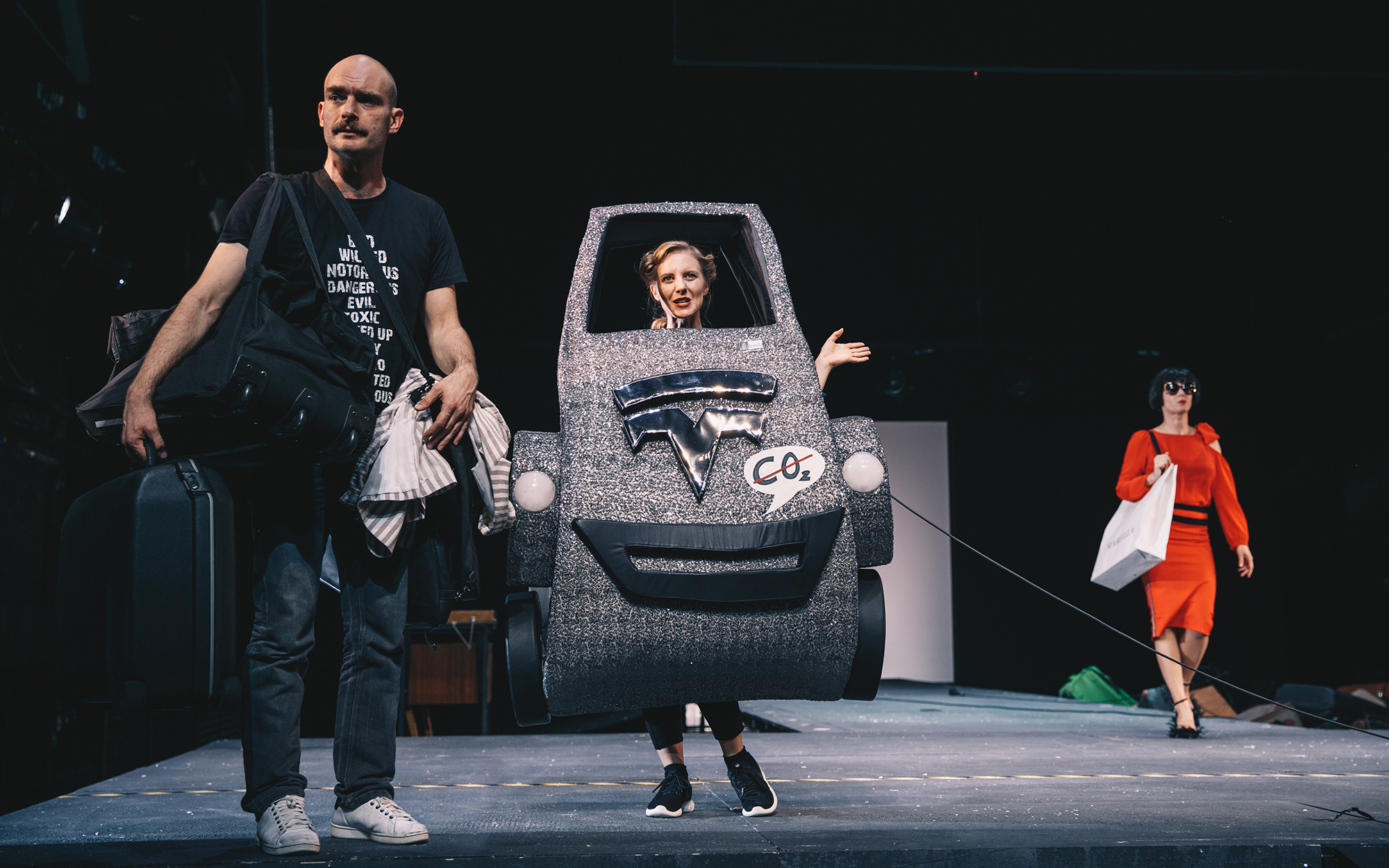 Toni (Gregor Gruden) je oprtan z vso svojo prtljago. Ob njem je Brenda (Lena Hribar) v maketi srebrnega avtomobila, ki dela reklamo za nizek izpust CO2. Iz ozadja v rdeči obleki prihaja Žana (Jana Zupančič) z nakupovalno vrečko na rami. Avtor fotografije: