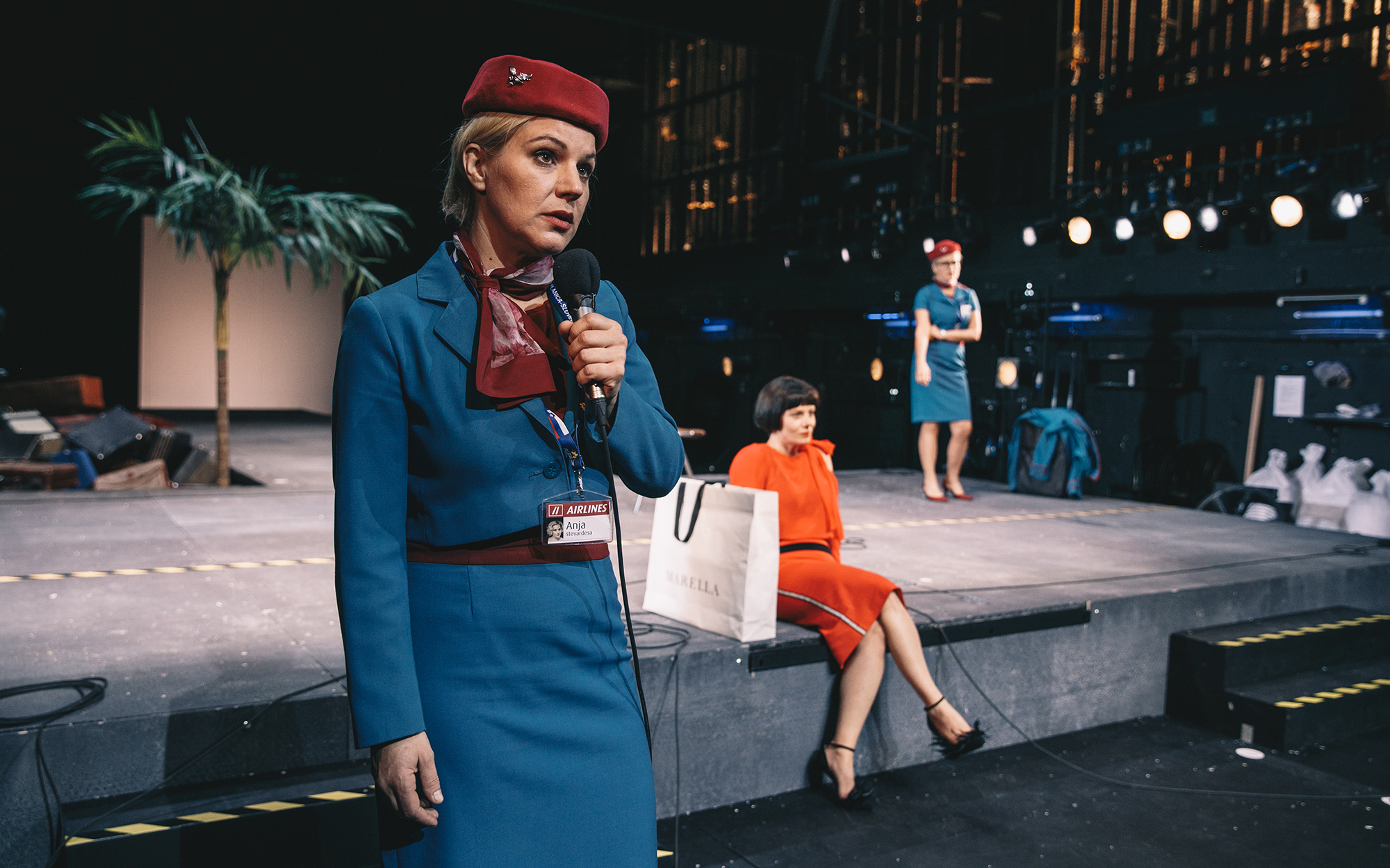 Stevardesi Anja (Karin Komljanec) in Lena (Tanja Dimitrievska) sta v enakih uniformah, z bordo klobučkom in ujemajočo se rutico. Anja (Karin Komljanec) govori v mikrofon, Lena (Tanja Dimitrievska) pa stoji v ozadju. Žana (Jana Zupančič) v rdeči obleki in 