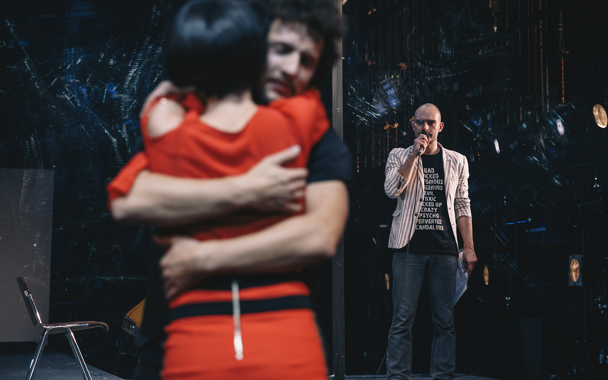 Toni (Gregor Gruden) za stekleno steno govori v mikrofon in opazuje objemajoča se Felipeja (Jernej Gašperin) in Žano (Jana Zupančič). Avtor fotografije: Peter Giodani.