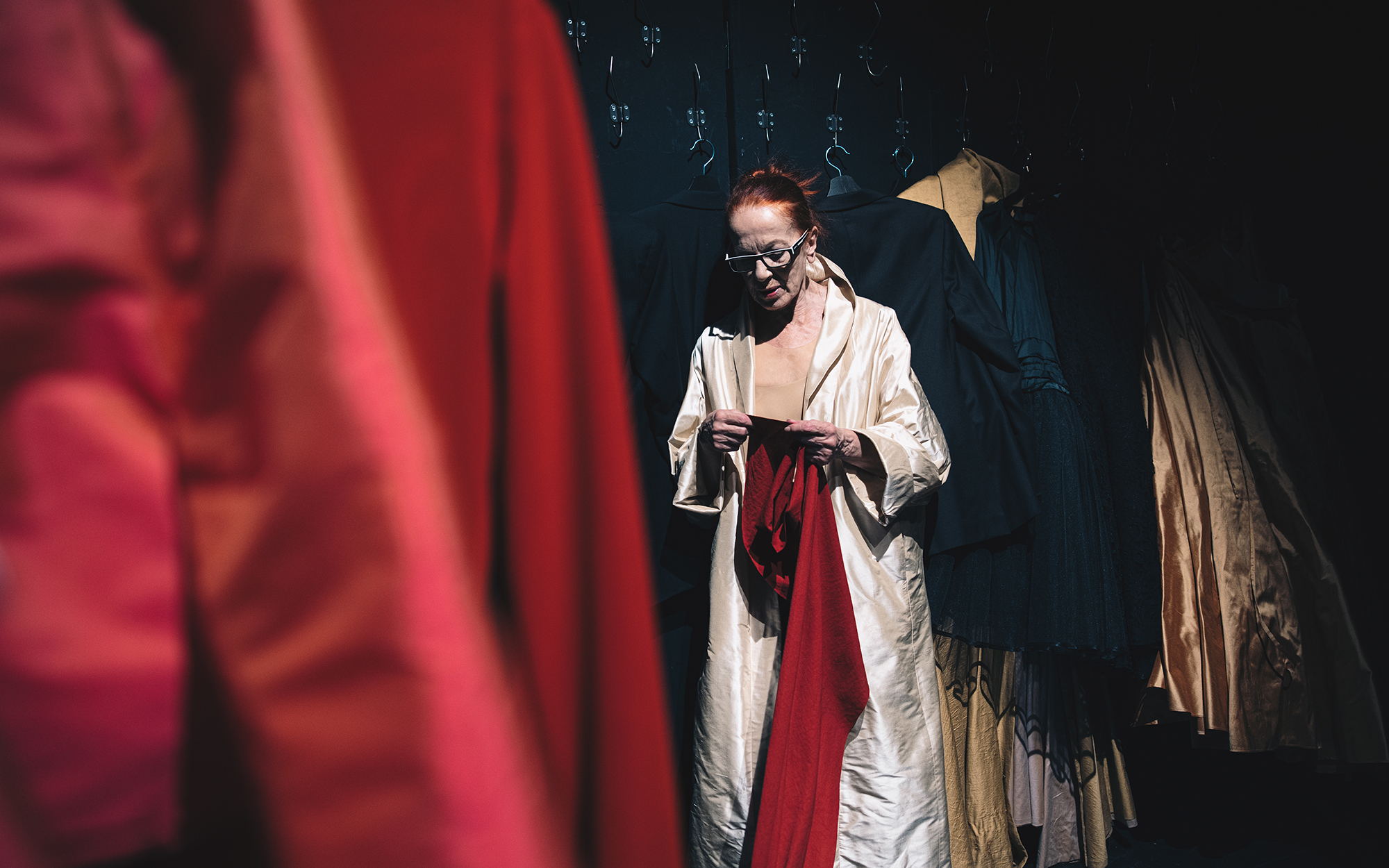 Jožica Avbelj stoji pred steno, na kateri je obešenih veliko najrazličnejših oblačil. Oblečena je v zlat plašč, v rokah zamišljeno drži dolgo rdečo tkanino.