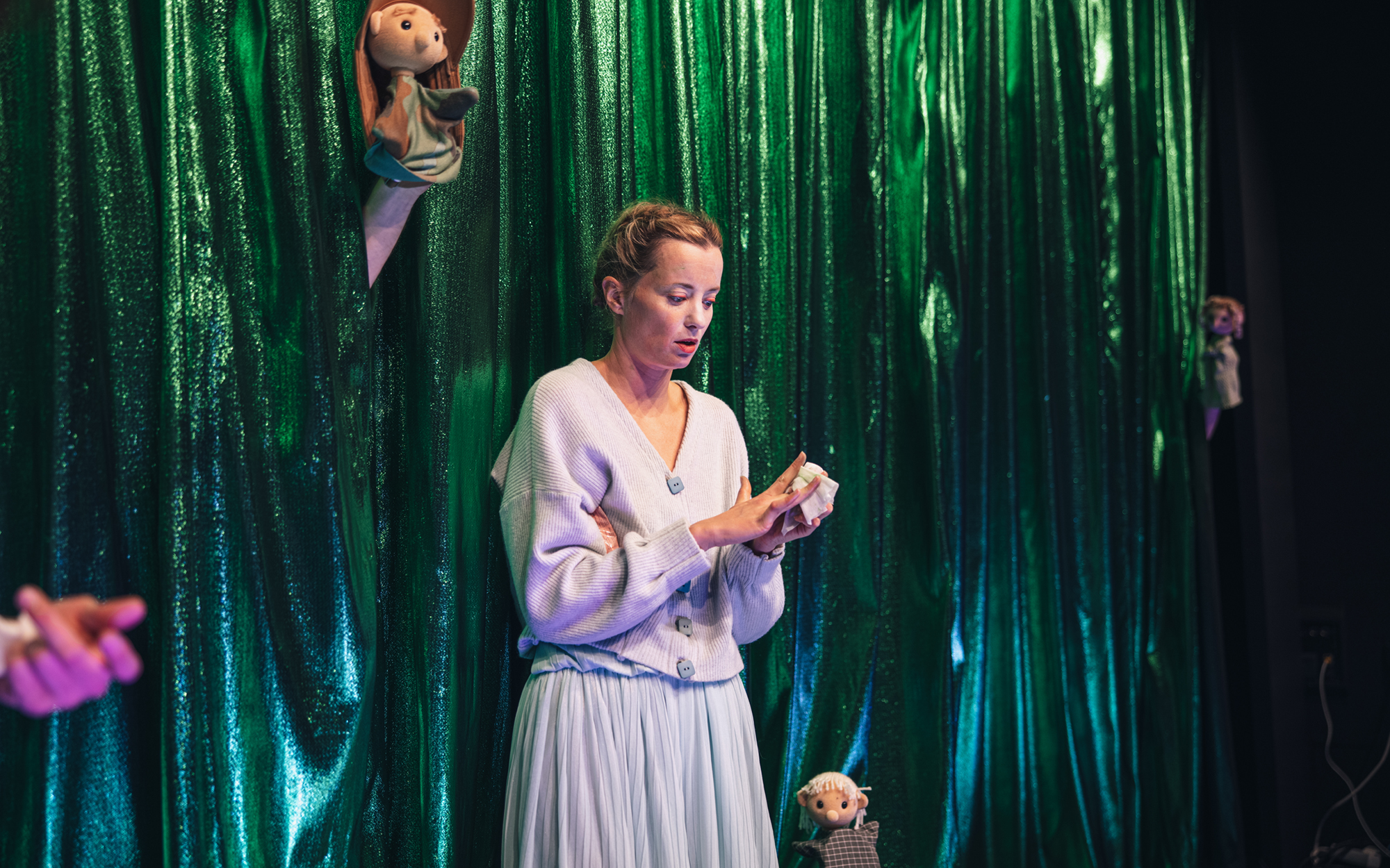 Ona (Ajda Smrekar) stoji pred bleščeče zeleno zaveso, z robčkom si briše roke, okoli nje izza zavese kukajo majhne lutke, ki spominjajo na Dobre ljudi. 