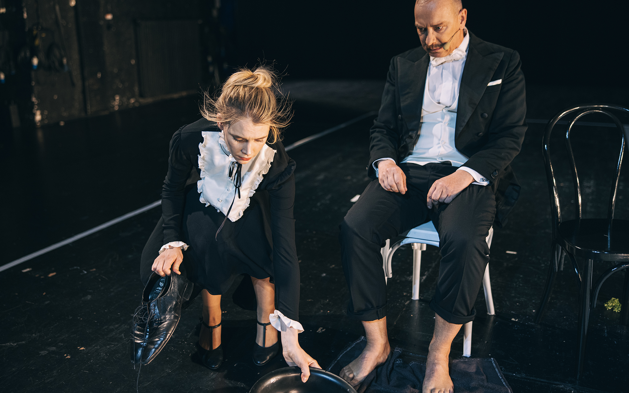 Virginija (Klara Kuk) čepi ob nogah Beauperthuisa (Uroš Smolej) in odmika lavor za umivanje nog, v eni roki drži par čevljev. Beauperthuis sedi na stolu, hlačnici ima zavihani in strmi predse. Avtor fotografije: Peter Giodani.