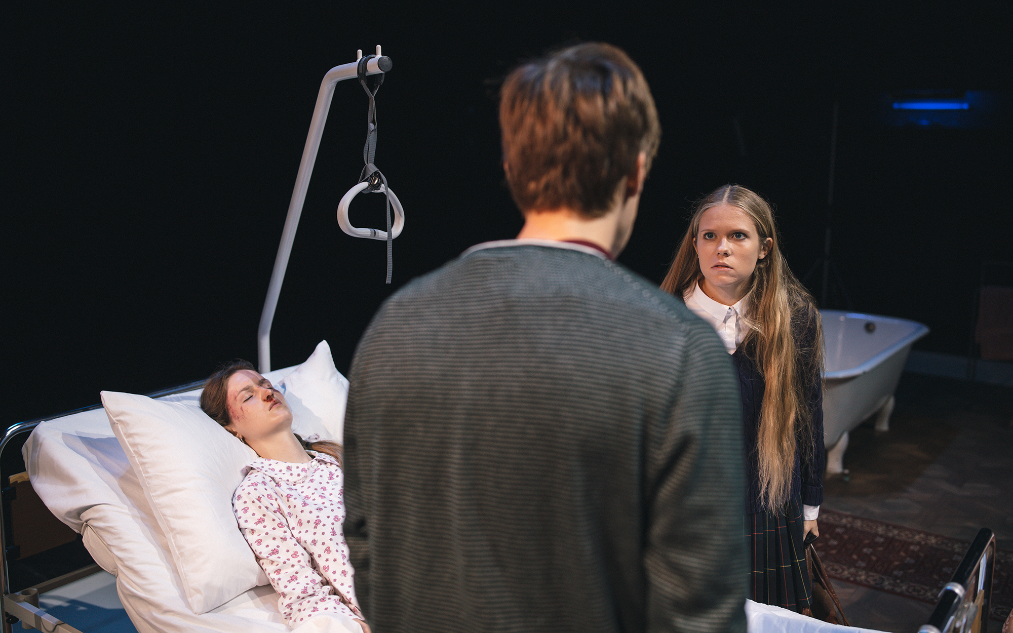Alice (Lucija Harum) leži na bolniški postelji z zaprtimi očmi in podplutbami na obrazu. Henry (Matej Zemljič) in Mia (Ana Pavlin) stojita vsak na eni strani postelje in se gledata. Avtor fotografije: Peter Giodani.