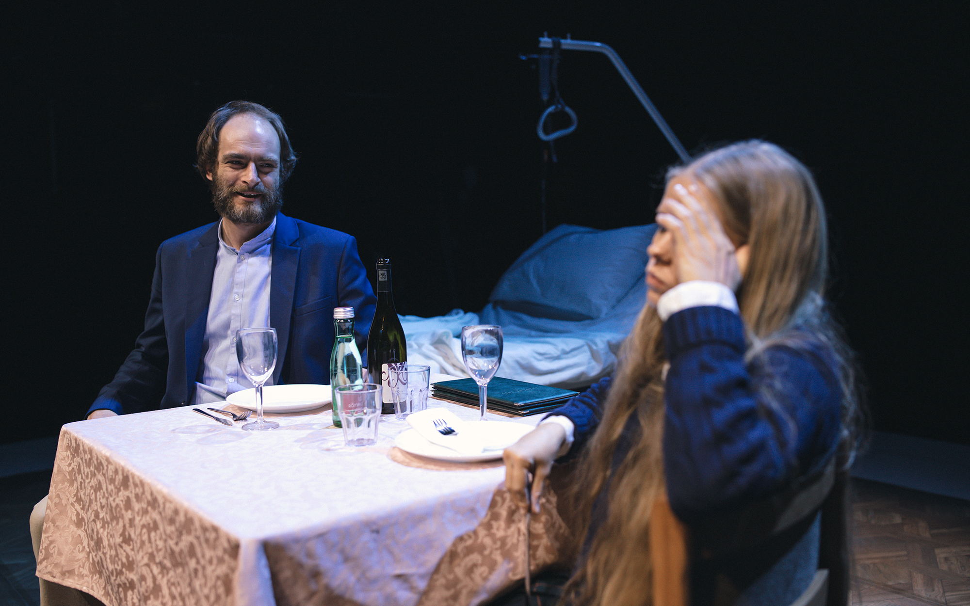 Hugh (Gregor Gruden) in Mia (Ana Pavlin) sedita v restavraciji ob pogrnjeni mizi. Ona se drži za glavo in se obrača stran od mize. Avtor fotografije: Peter Giodani.