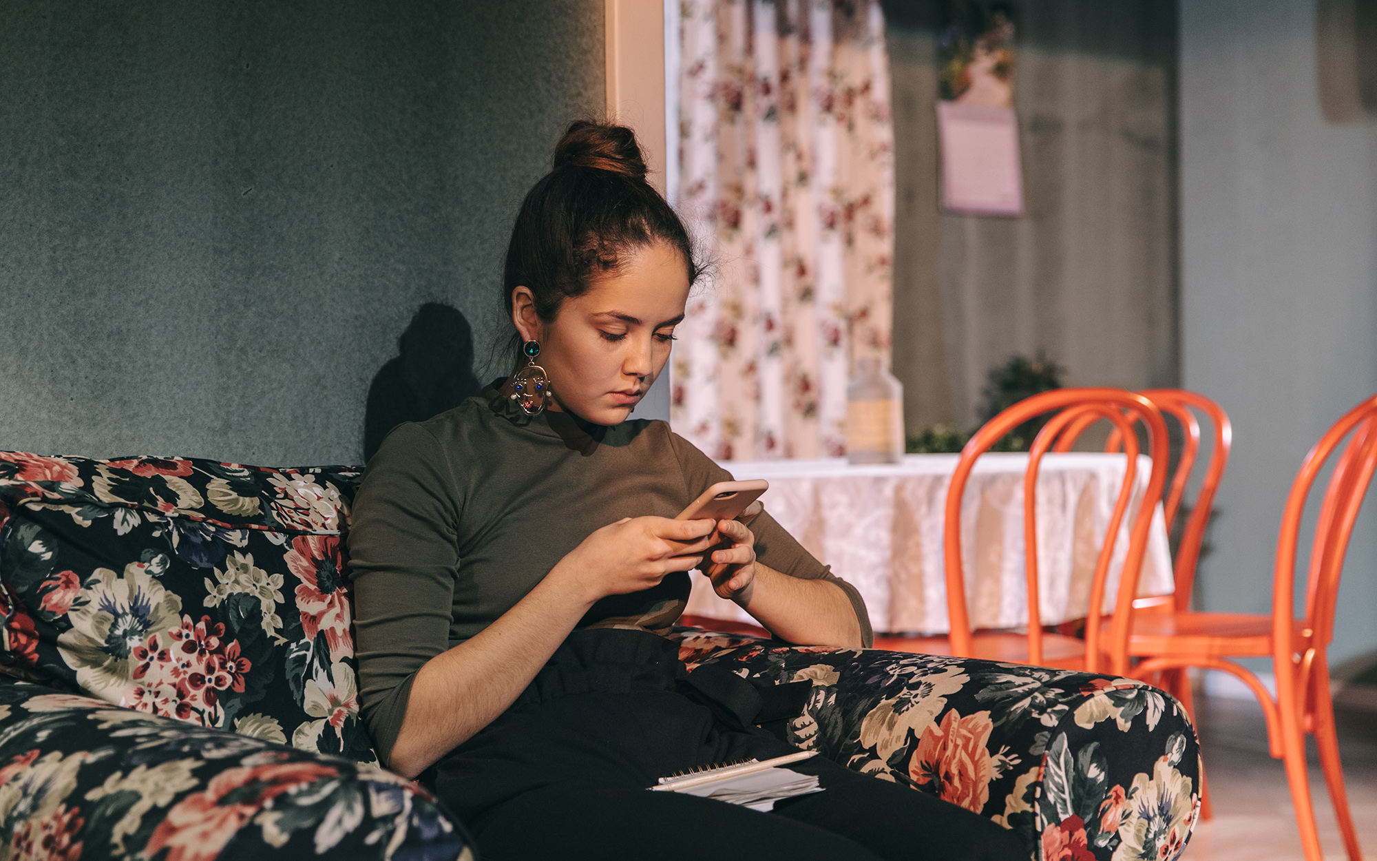 Tamala (Lara Wolf) sedi v naslanjaču in gleda v mobilni telefon. Avtor fotografije: Peter Giodani.