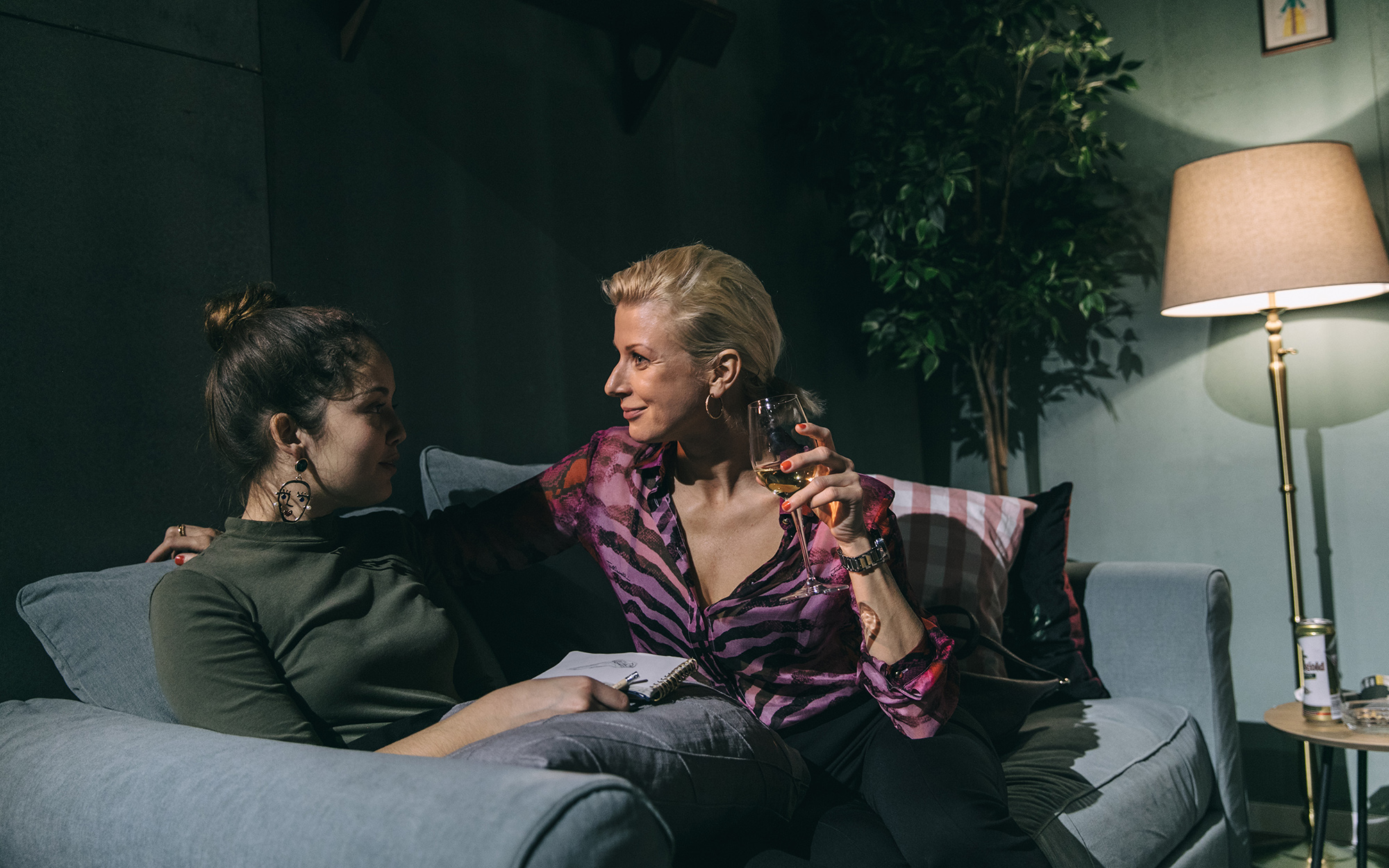 Tamala (Lara Wolf) in Maja (Tjaša Železnik) sedita na kavču v dnevni sobi in se pogovarjata. Tamala ima v rokah skicirko in svinčnik, Maja pa kozarec oranžnega vina. Avtor fotografije: Peter Giodani.