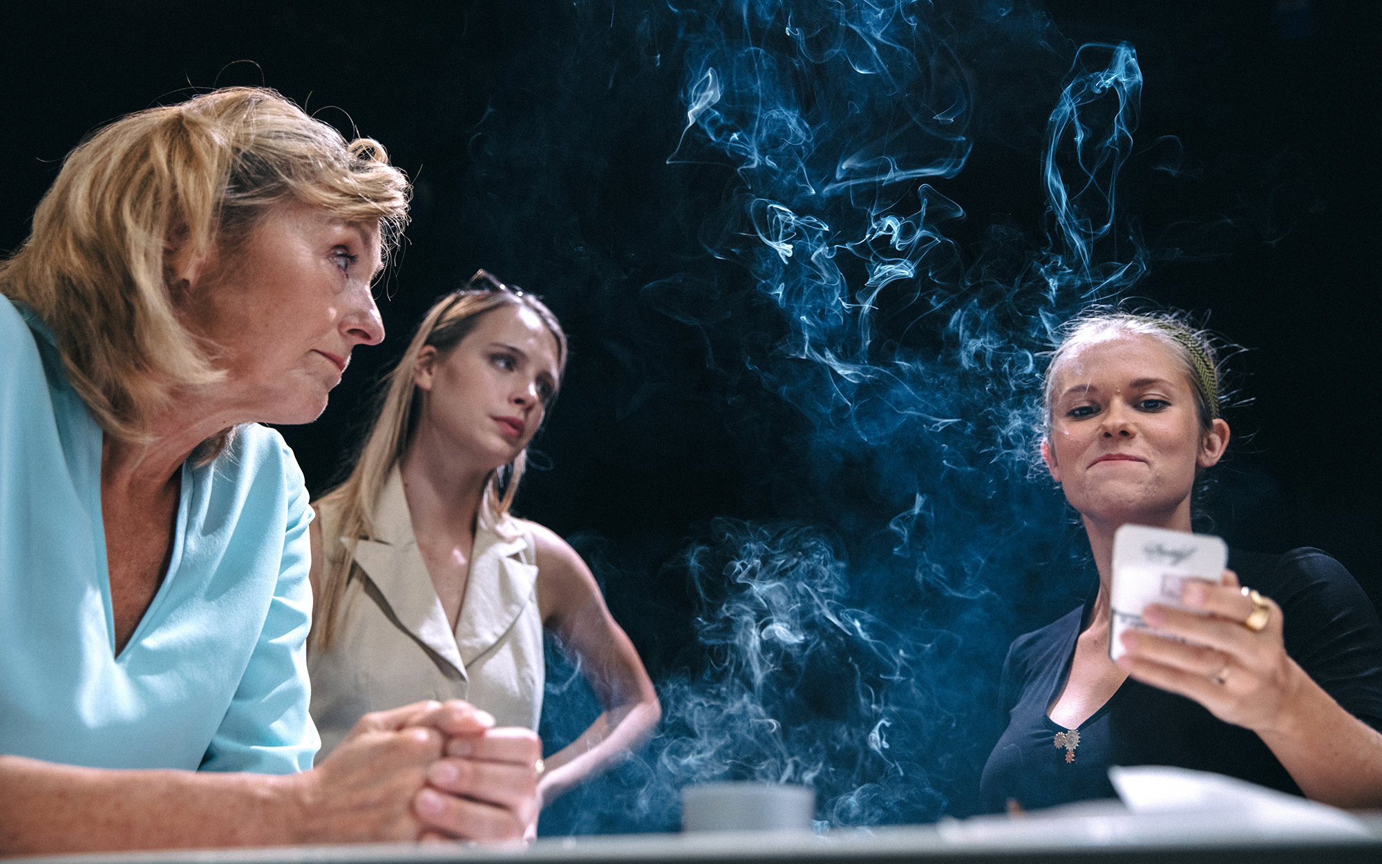 Hana (Judita Zidar) in Karla (Klara Kuk) opazujeta Lizo (Ana Pavlin), ki se premeteno nasmiha. Obdaja jo cigaretni dim.  Avtor fotografije: Peter Giodani.