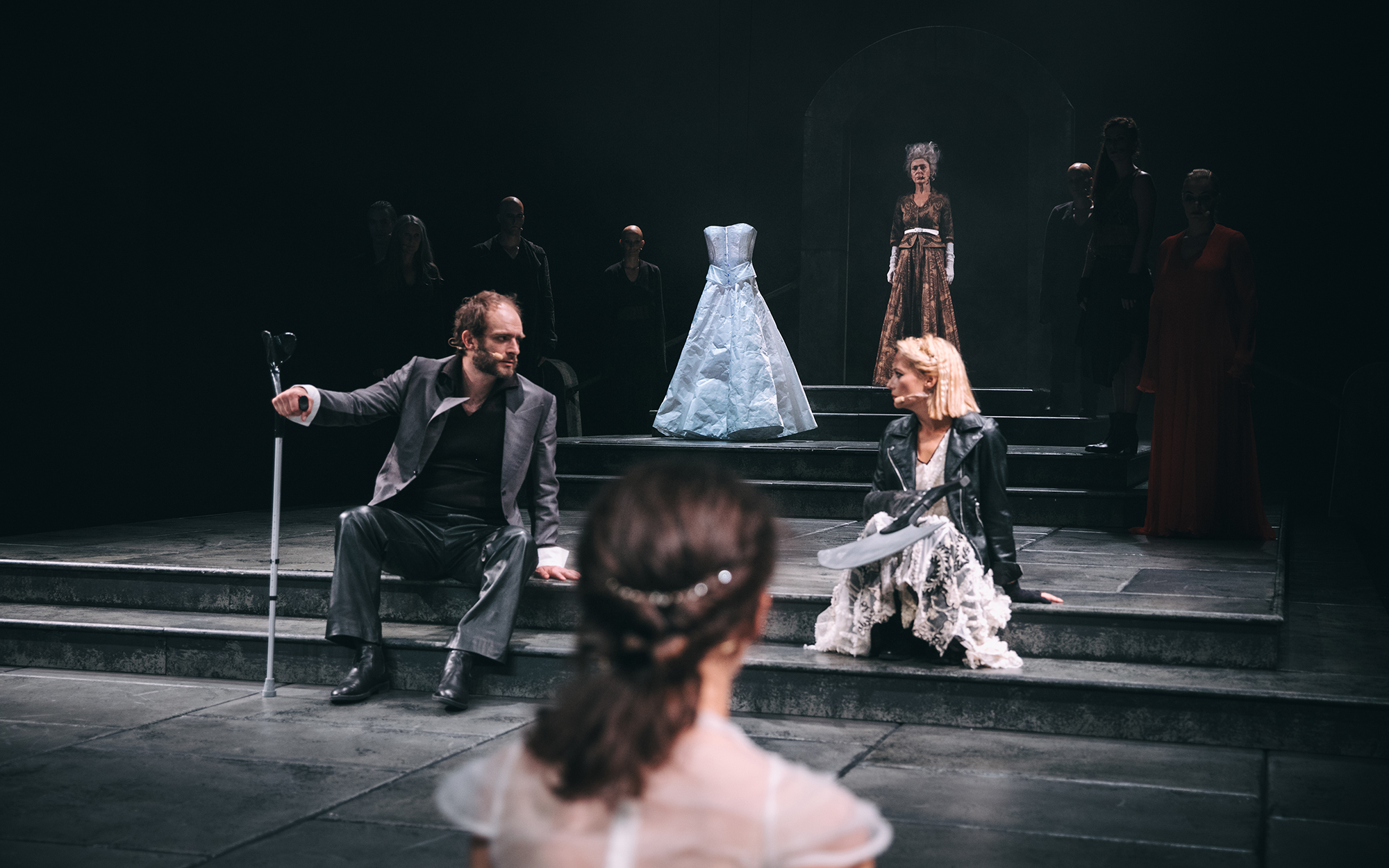 Kalaf (Gregor Gruden) sedi na stopnicah in se obrača k Turandot (Viktorija Bencik Emeršič), ki drži v naročju rabljevo sekiro. V ozadju Kraljica mati (Mirjam Korbar) in Mlada Turandot (Lara Wolf). Na stopnicah stojalo z belo poročno obleko. Avtor fotograf