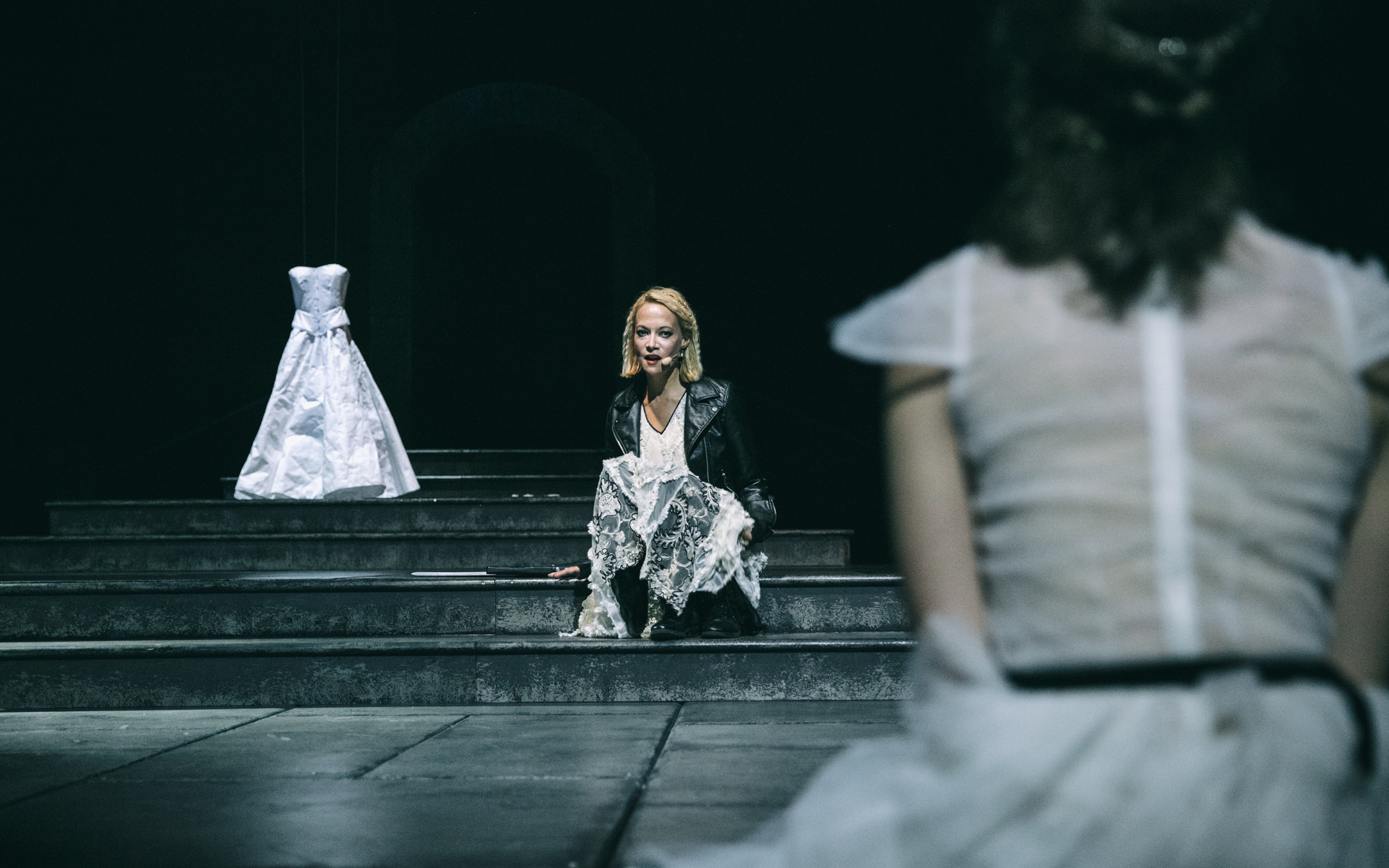 Turandot (Viktorija Bencik Emeršič) sedi na stopnicah in se obrača k Mladi Turandot (Lara Wolf). V ozadju bela poročna obleka. Avtor fotografije: Peter Giodani.