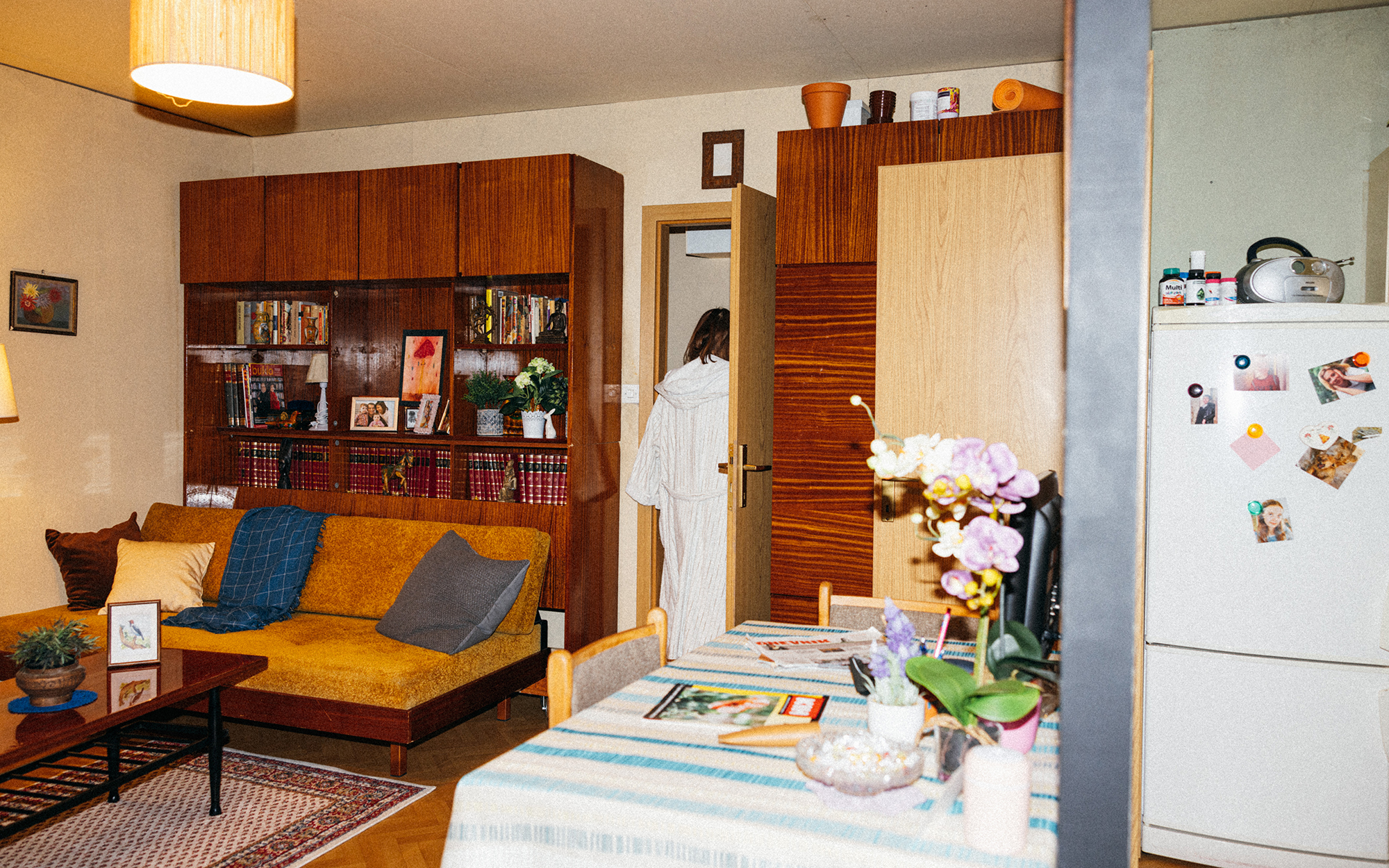 Mama (Mirjam Korbar) v beli jutranji halji odhaja iz dnevne sobe na hodnik. Dnevna soba je lepo pospravljena. Na levi strani na steni visi uokvirjena slika z motivom šopka, pod sliko je kavč s tremi blazinami različnih barv, čezenj je pogrnjena modra odej